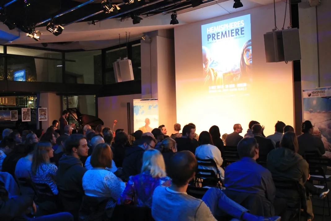 Buchpremiere in einem Saal: Publikum vor einer großen Projektion mit dem Plakat „Hemispheres Premiere“.