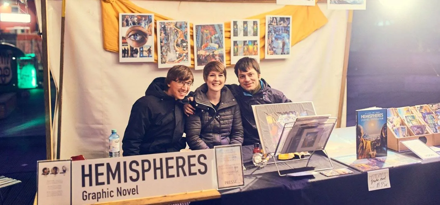 Drei Teammitglieder sitzen hinter dem Messestand von Hemispheres, hinter ihnen hängen ausgedruckte Comicseiten, davor liegen Bücher und Postkarten.